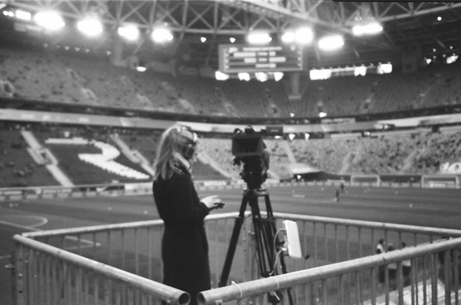 Black and white side view of female camera operator with blonde hair in black coat and headsets standing in front of professional video camera at stadium