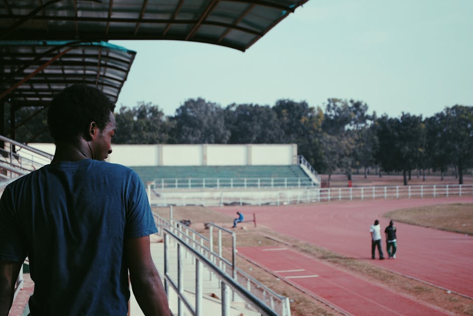 A man stands in a stadium, overlooking a track with athletes practicing in the distance.