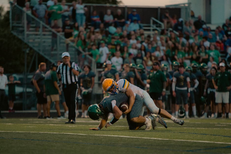 Two football players in intense tackle, surrounded by excited crowd during a night game.